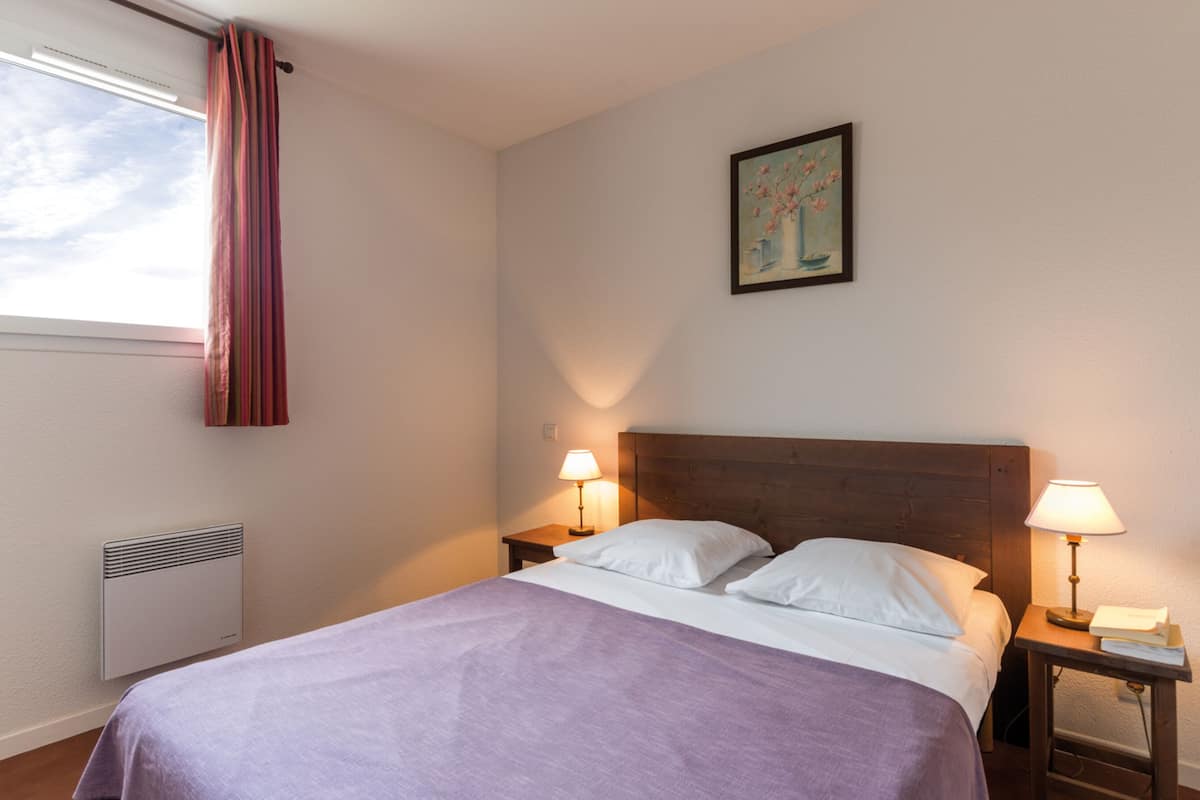 A neatly arranged hotel room with a bed, two bedside lamps, a radiator, a window with curtains, and a painting on the wall.
