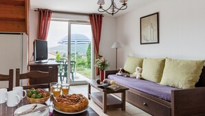 A cozy living room with a sofa, a dining table with breakfast items, a TV, and a view of the outdoors. - Le Domaine des Cazelles by Ateya Vacances (Cajarc)