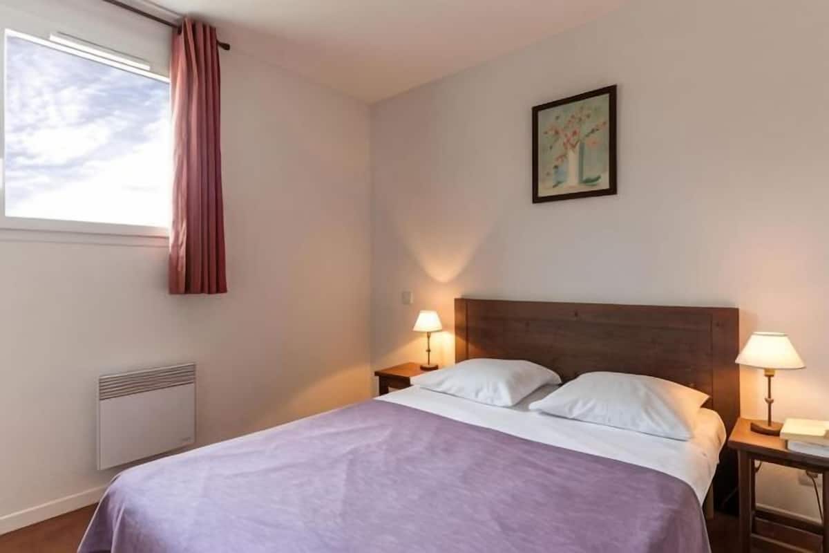 A hotel room with a bed, two bedside lamps, a window with curtains, a radiator, and a painting on the wall.