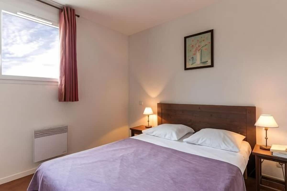 A bedroom with a bed, two bedside tables with lamps, a window with a curtain, a radiator, and a painting on the wall.