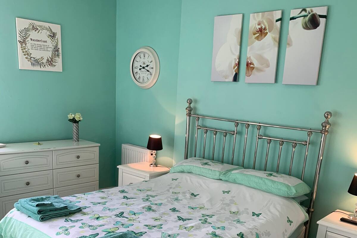 Ein Schlafzimmer mit Bett, Nachttischen, einem Schminktisch, einer Uhr und Wandbildern.