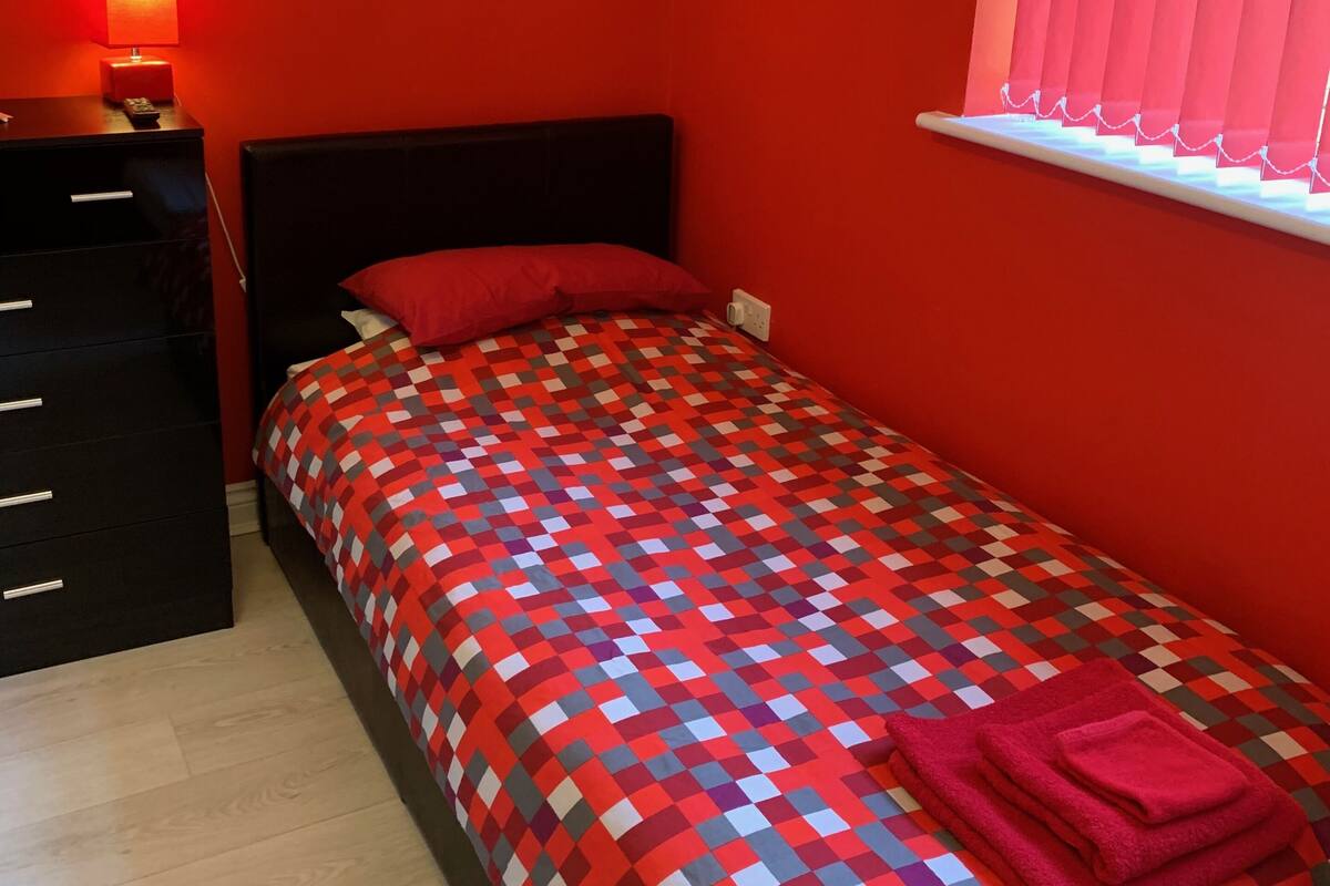 Ein Schlafzimmer mit Bett, Nachttisch, Lampe, einem Gemälde und einem Fenster mit Vorhängen.