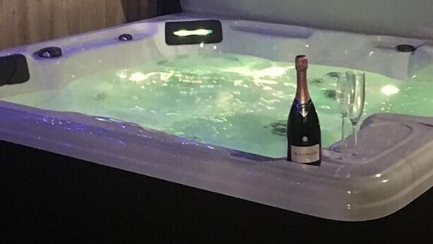 A hot tub with a bottle of champagne and a glass, surrounded by wooden walls and string lights. - BedrockLodge Buisness Centre HolidayLets (Bristol)