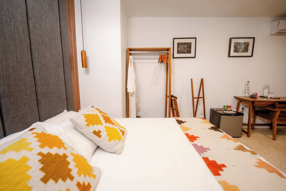 Ein Hotelzimmer mit einem Bett, einem Schreibtisch und einem Stuhl. Es gibt einen Kleiderschrank, einen Kleiderhaken und einen kleinen Tisch mit einer Flasche und einer Tasse.