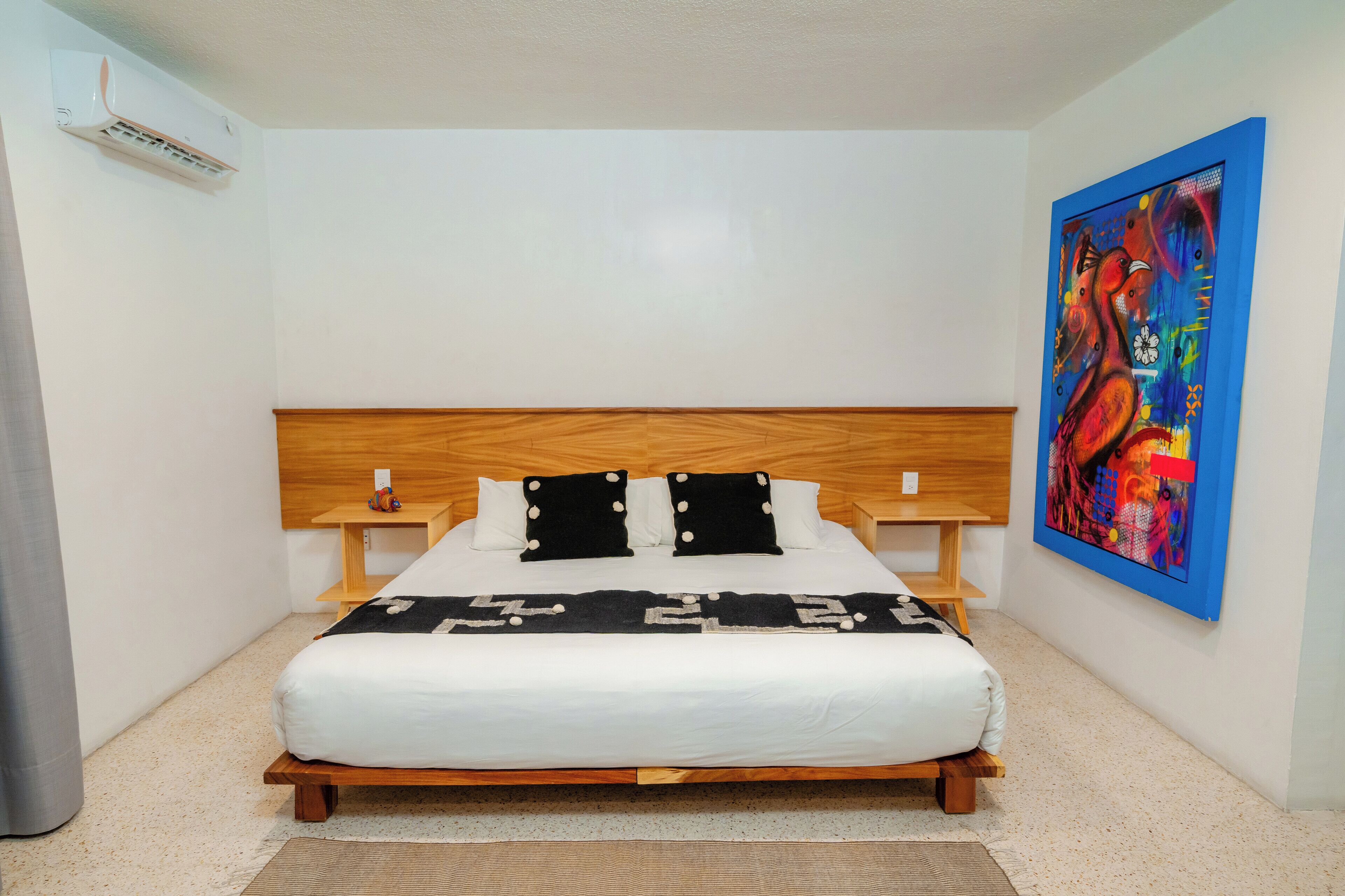 A neatly made bed with a wooden headboard, a bedside table with a lamp, and a colorful abstract painting on the wall.