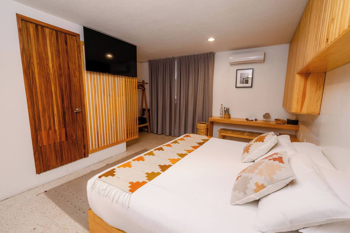 Ein Schlafzimmer mit einem Bett, einem Kleiderschrank, einem Fernseher, einem Fenster mit Vorhängen und einem kleinen Tisch mit Gegenständen darauf.