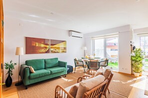 A modern living room with a green sofa, a dining area with a table and chairs, and a large window with a view of the outside. - ShôLubi - Charming Quiet & Cozy (Lisbon)
