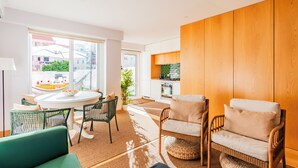 A modern living room with a green sofa, wicker chairs, a round dining table, and a view of the city through large windows. - ShôLubi - Charming Quiet & Cozy (Lisbon)