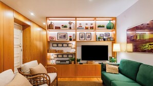 A cozy living room with a green sofa, a wooden TV stand with shelves, a wicker chair, and a flat-screen TV. - ShôLubi - Charming Quiet & Cozy (Lisbon)