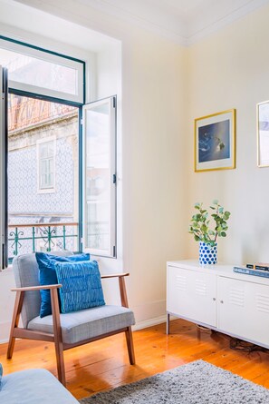 A room with a wooden floor, a grey armchair with blue cushions, a white cabinet, and a window with an open shutter. - My Graça Flat in Lisbon (Lisbon)