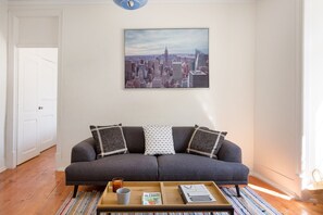 Un salon avec un canapé gris, des coussins à motifs, une table basse avec des magazines et un tableau représentant un paysage urbain accroché au mur.