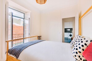 Une chambre à coucher avec un lit, une fenêtre donnant sur un bâtiment à toit rouge, une cuisine dans la pièce adjacente et une chaise en bois.