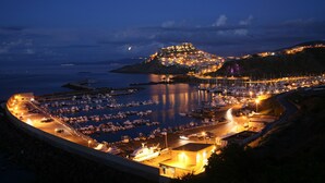 A coastal marina at night with boats docked, a prominent hilltop structure, and a curved road leading to the marina. - Affittimoderni Castelsardo Lentisco - Csle03 (Castelsardo)