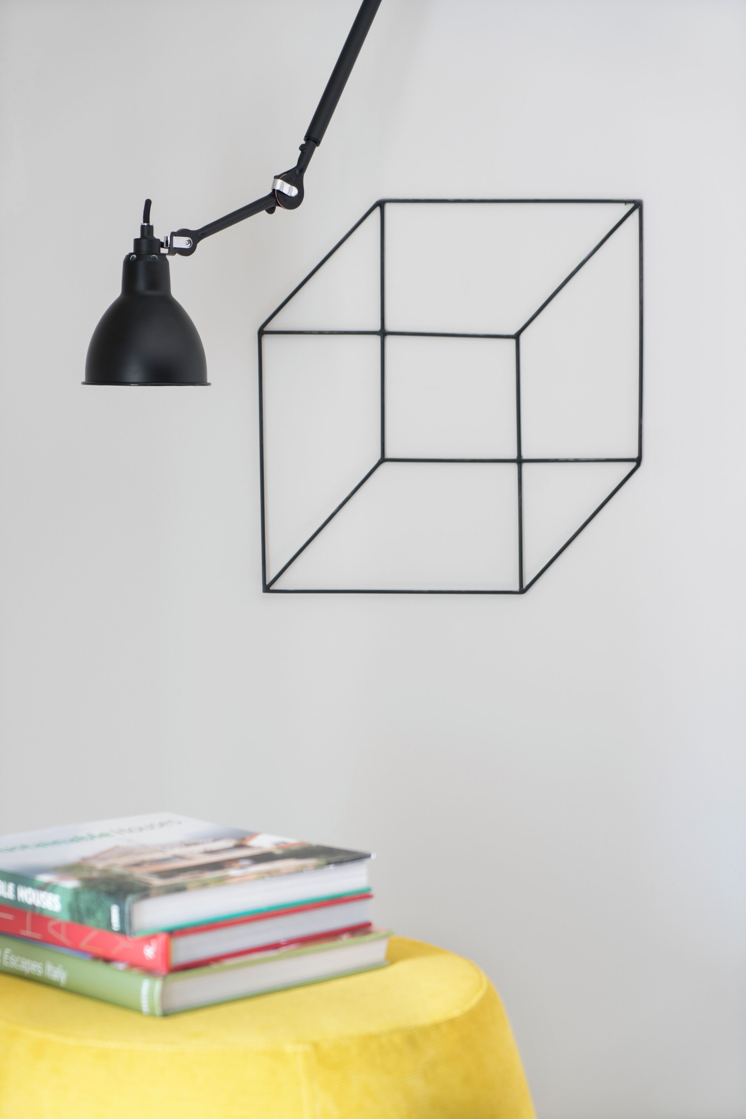 A wall-mounted geometric art piece, a black desk lamp, and a stack of books on a yellow ottoman.