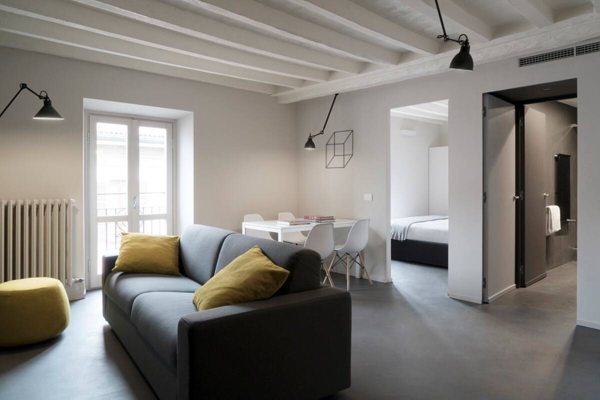Ein modernes Wohnzimmer mit grauem Sofa, gelben Kissen, einem Essbereich mit weißem Tisch und Stühlen sowie einer Küchenzeile mit Kühlschrank.