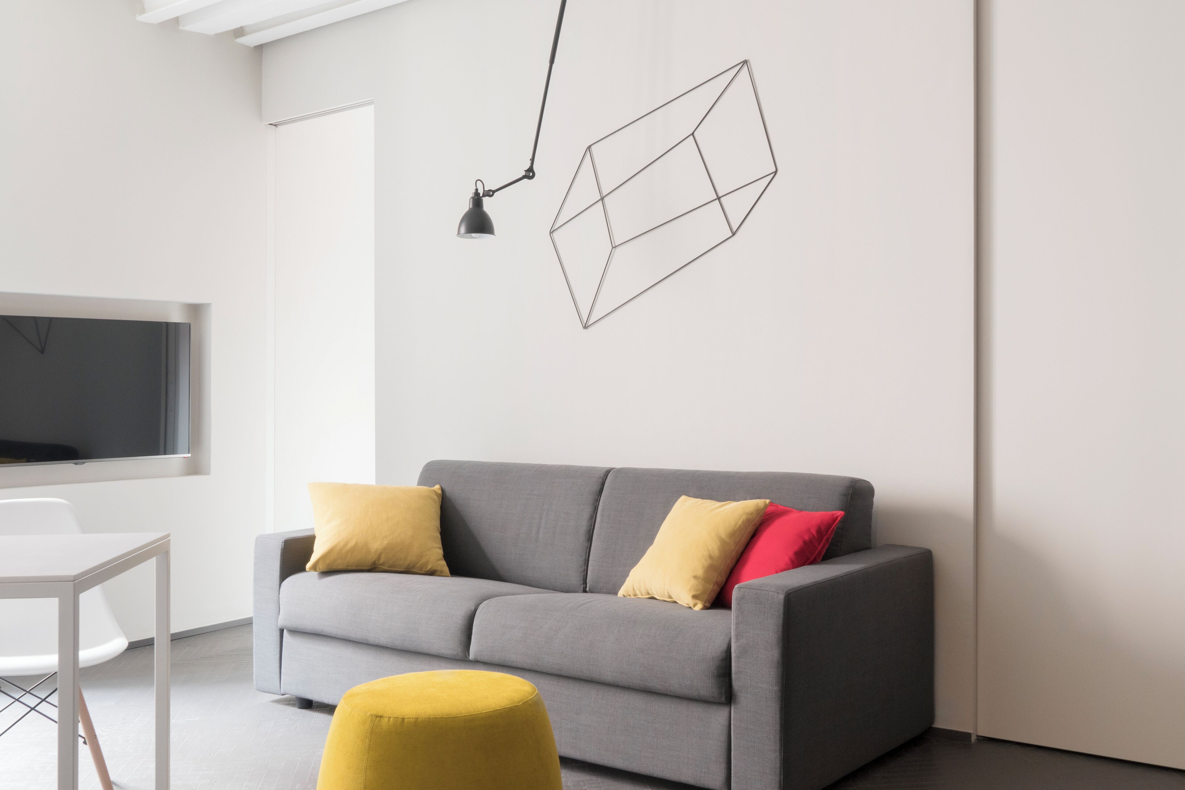 A modern living room with a grey sofa, yellow and red cushions, a yellow ottoman, a white table, and a geometric wall art piece.