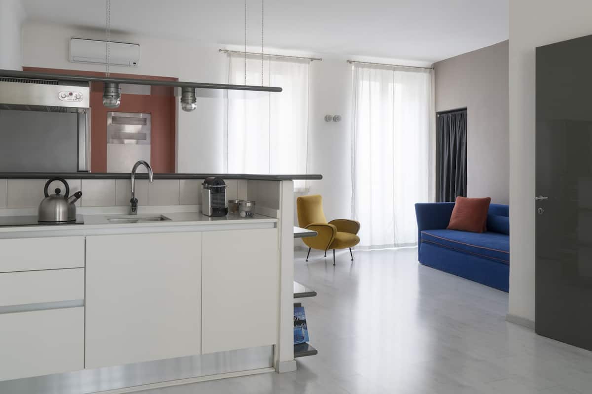Eine moderne Küche mit Einbauofen, Spüle und Wasserkocher. Im Wohnbereich befindet sich ein blaues Sofa mit einem roten Kissen.
