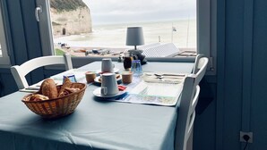 Tavola per la colazione con un cestino di pane, una tazza e una mappa, con vista su una spiaggia, barche e scogliere.
