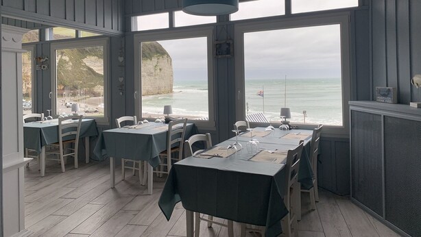 Un ristorante con tavoli apparecchiati per la cena, ampie finestre che offrono vista sul mare e sulle scogliere, e un lampadario.