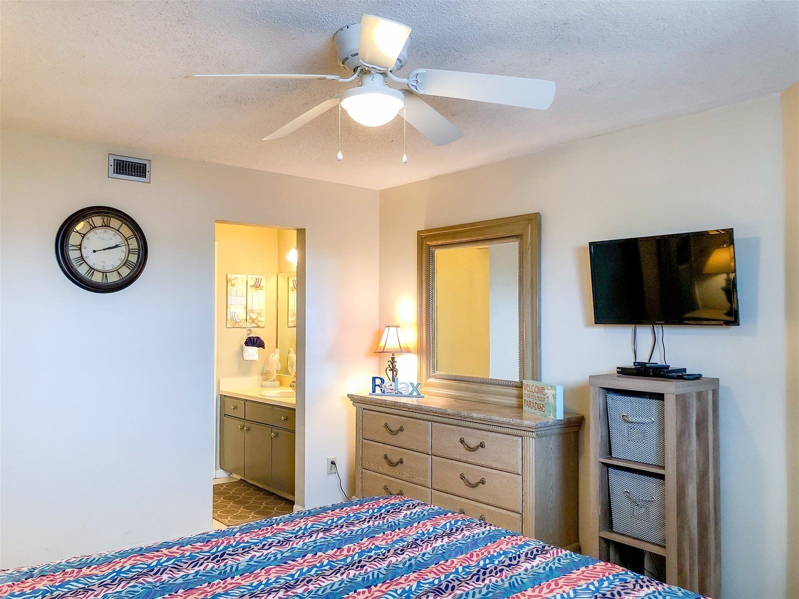 Une chambre à coucher comprenant un lit, un ventilateur de plafond, une horloge, un miroir, un téléviseur et une commode.