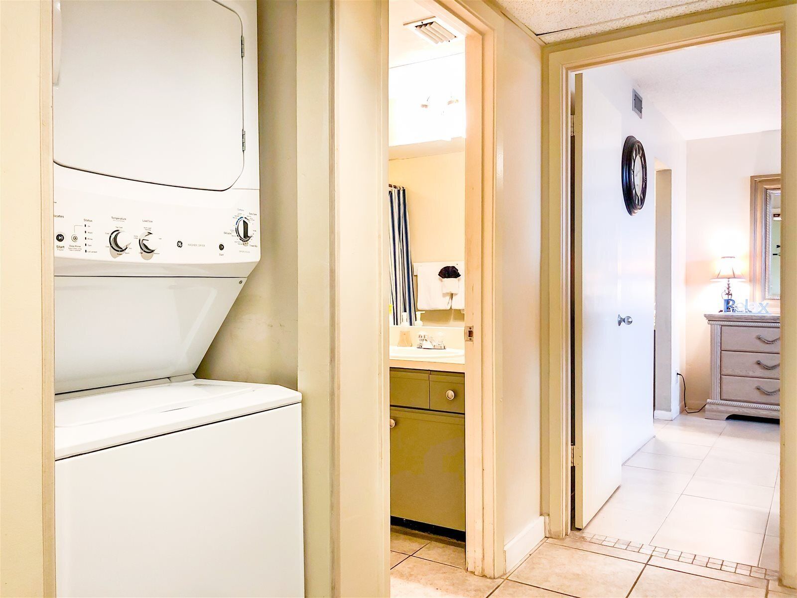 A laundry area with a washing machine and dryer, a bathroom with a sink and mirror, and a hallway leading to a bedroom with a dresser and lamp.