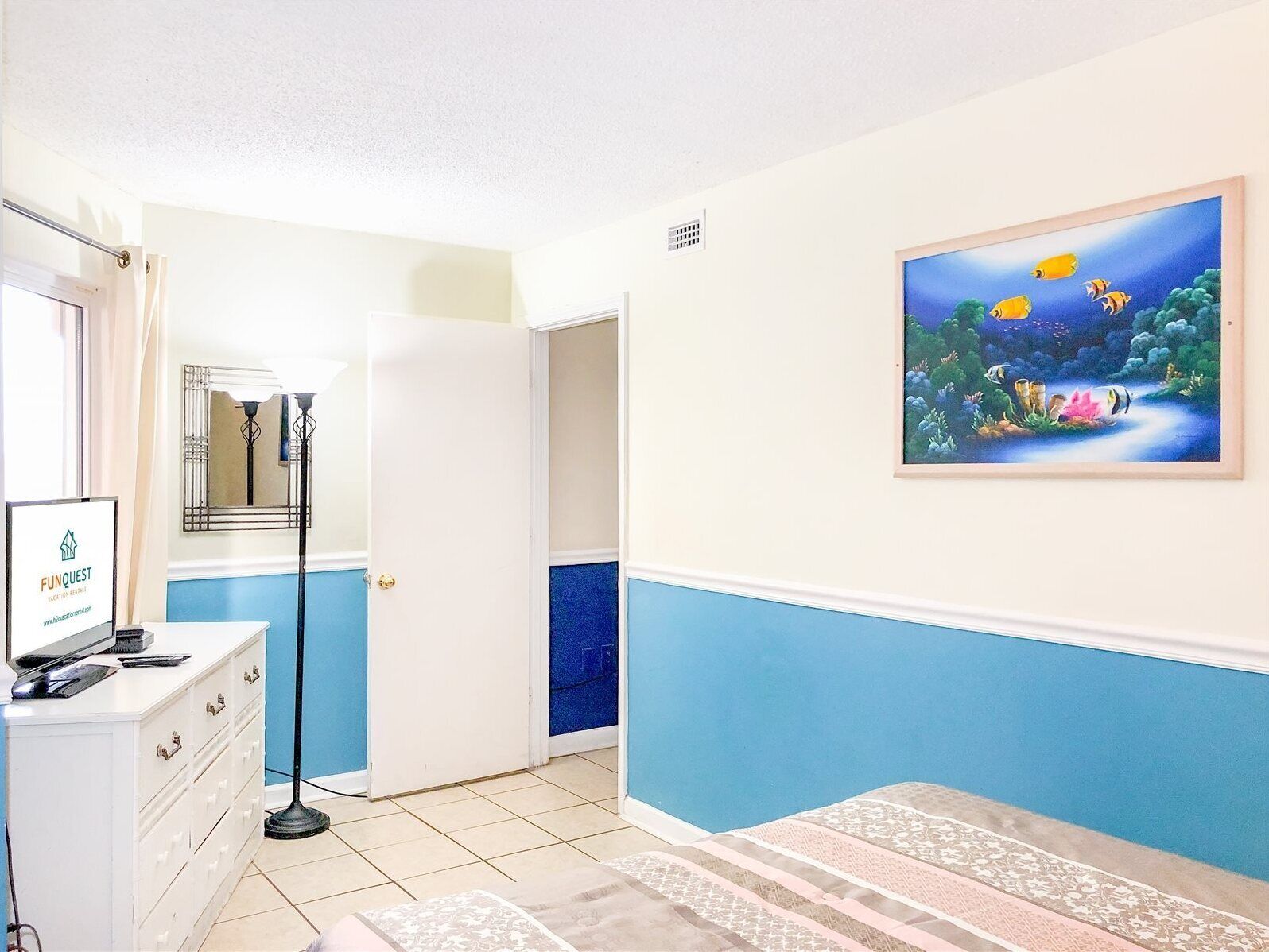 A hotel room with a bed, a desk with a computer, a mirror, and a painting of fish on the wall.