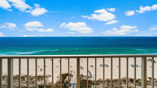 Ein Strand mit blauem Himmel und Meer. Menschen liegen am Strand, im Vordergrund ist ein Geländer zu sehen.
