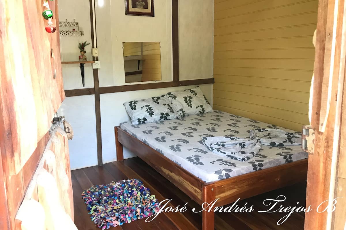 Une pièce avec un lit, un plancher en bois, un miroir et un ventilateur fixé au mur.