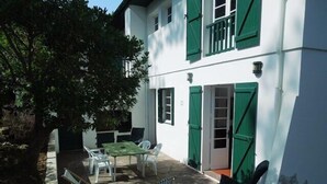 A white building with green shutters, a patio with a table and chairs, and a tree.