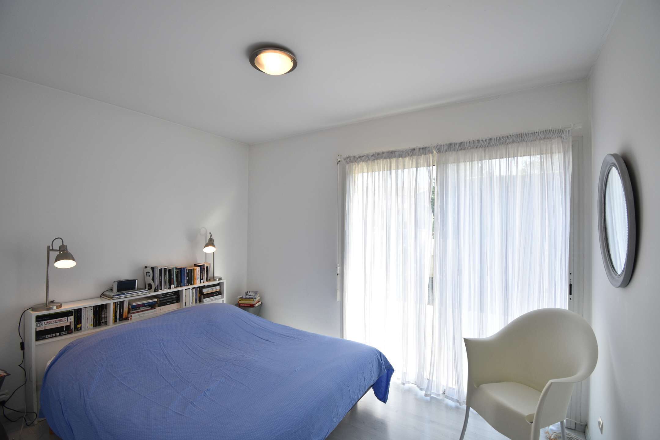 A bedroom with a bed, a chair, a desk with books and a computer, and a window with curtains.