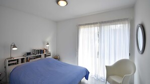 Une chambre à coucher avec un lit, une chaise, un bureau avec des livres et un ordinateur, et une fenêtre avec des rideaux.