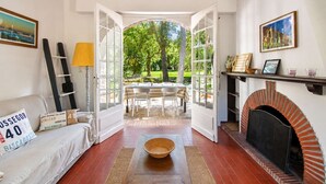 Une pièce comprenant un canapé, des portes ouvertes donnant sur un jardin, une cheminée et un coin repas.