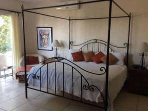 Uma cama de dossel com roupa de cama branca e várias almofadas, uma pequena mesa de cabeceira com abajur e uma obra de arte emoldurada na parede.