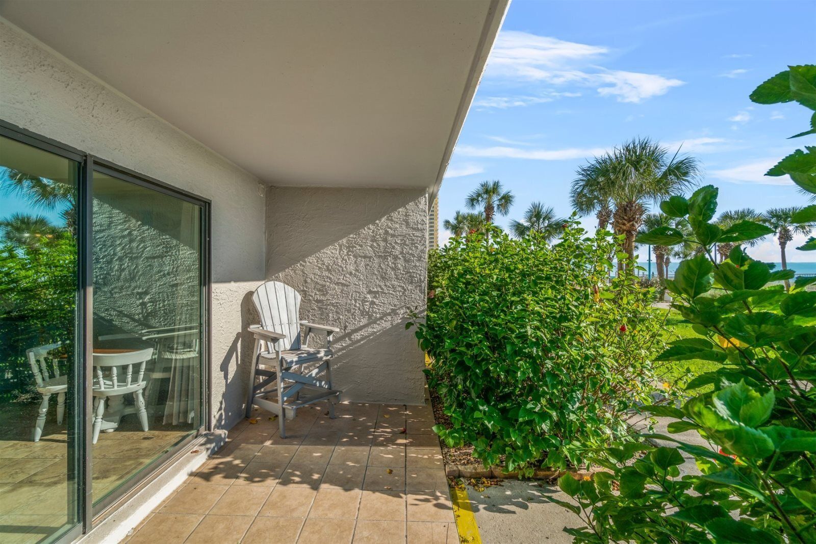A balcony with a white Adirondack chair, a glass door, and a view of palm trees and the ocean.