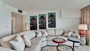 Een moderne woonkamer met een beige hoekbank, drie schilderijen van zeepaardjes, een lamp en twee bijzettafels met decoratieve objecten.