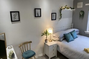 A bedroom with a bed, bedside table, chair, and wall-mounted shelves with plants. - 17G Regent Place (Clydebank)