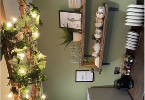 L’intérieur d’un café avec un mur vert, une enseigne représentant une tasse de café, des étagères remplies de tasses et de verres, et une plante décorée de guirlandes lumineuses.