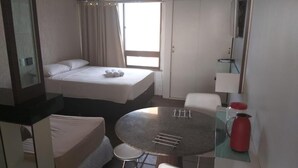 A compact hotel room with a bed, a small round table, a chair, a red kettle, and a window with curtains. - Flat Frente para o Mar Praia de Iracema (Fortaleza)