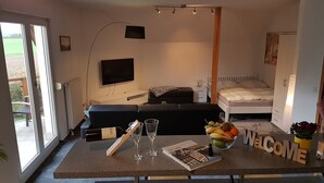 Quarto de hotel moderno com sofá-cama, TV de tela plana, mesa de cabeceira pequena, abajur, cesta de frutas e placa de boas-vindas.