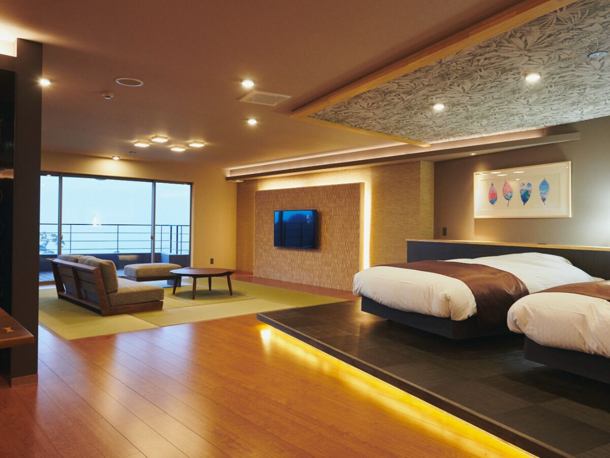A modern hotel room with a large bed, a seating area with a sofa and armchair, a flat-screen TV mounted on the wall, and a balcony with a view.
