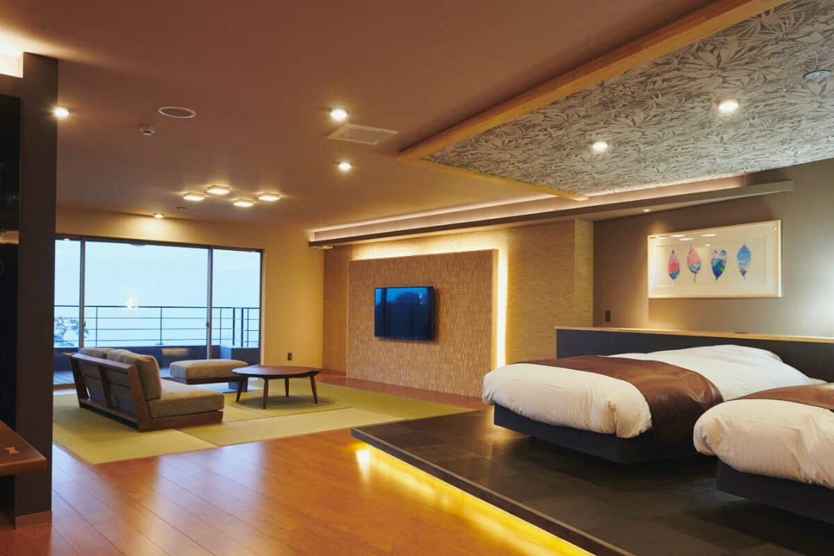 A modern hotel room with a large bed, a seating area with a sofa and armchair, a flat-screen TV mounted on the wall, and a balcony with a view.