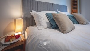 Un lit bien fait, agrémenté d’oreillers blancs et turquoise, une lampe de chevet et une assiette de croissants et de fraises posée sur une table de chevet.