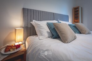 A neatly made bed with white and teal pillows, a bedside lamp, and a plate of croissants and strawberries on a nightstand.