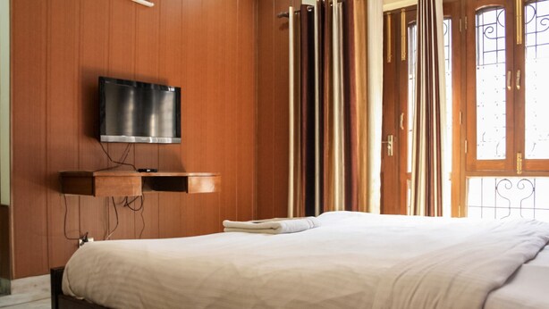 Een slaapkamer met een bed, een flatscreen televisie bevestigd aan een houten paneel, een nachtkastje en een raam met gordijnen.