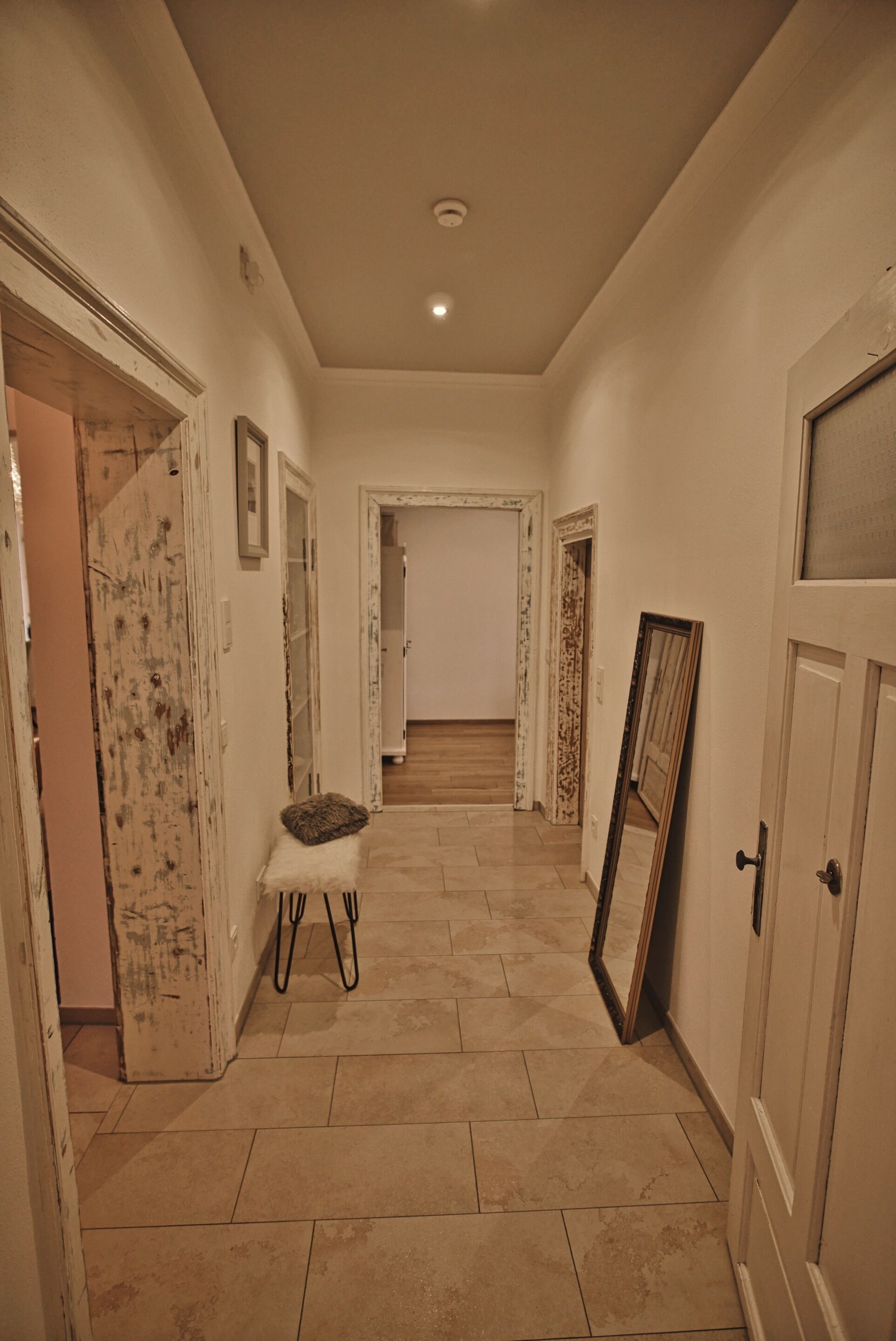 Un couloir avec un banc, un miroir appuyé contre un mur et une porte ornée d’un cadre décoratif.
