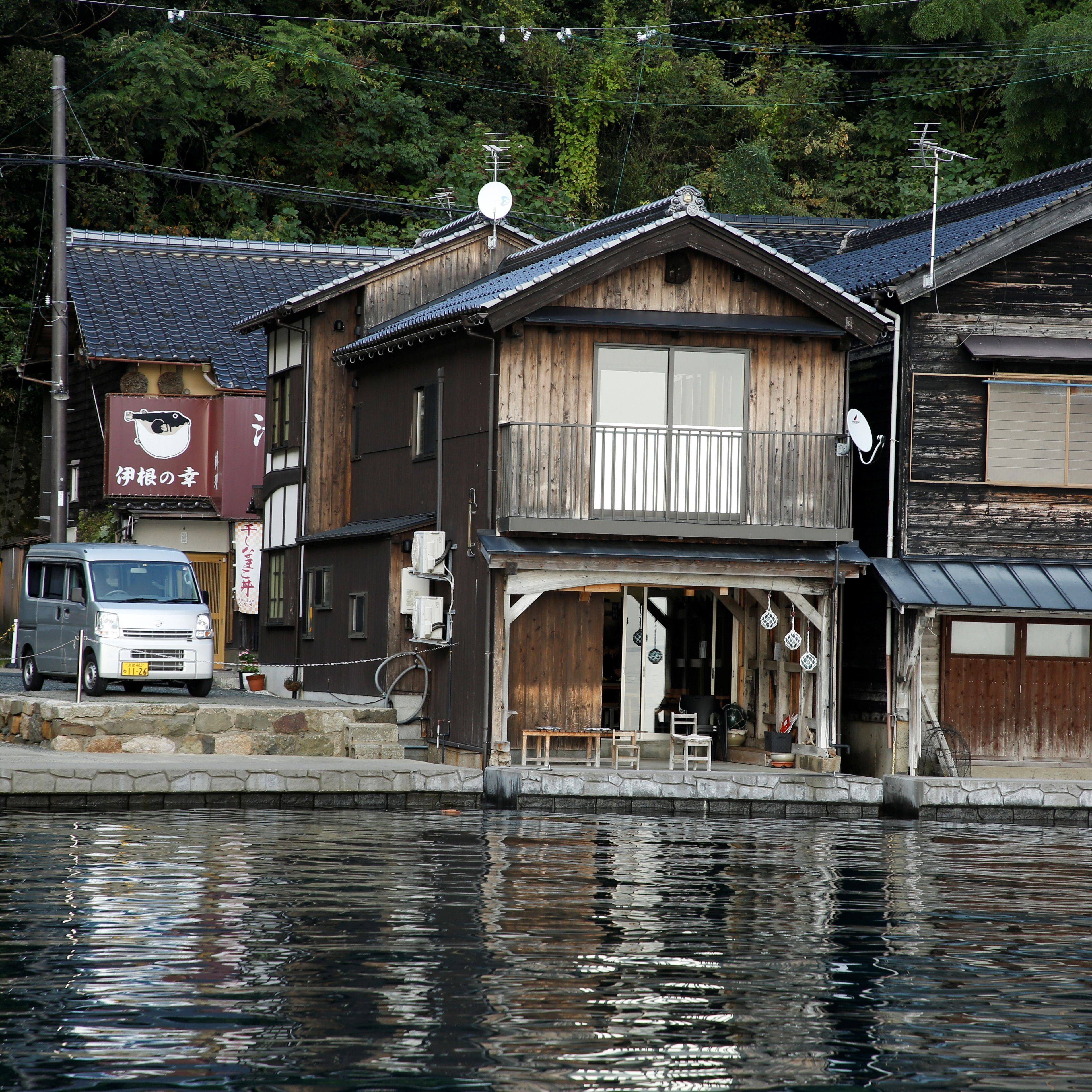 A traditional wooden building with a balcony, a van parked in front, and a river in the foreground.