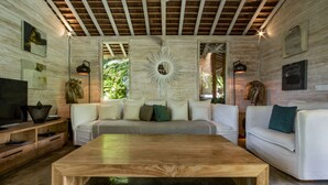 Amplia sala de estar con una gran mesa de centro de madera, un sofá y un sillón blanco. Hay un televisor sobre un soporte de madera, y la habitación cuenta con un techo de madera con vigas a la vista.