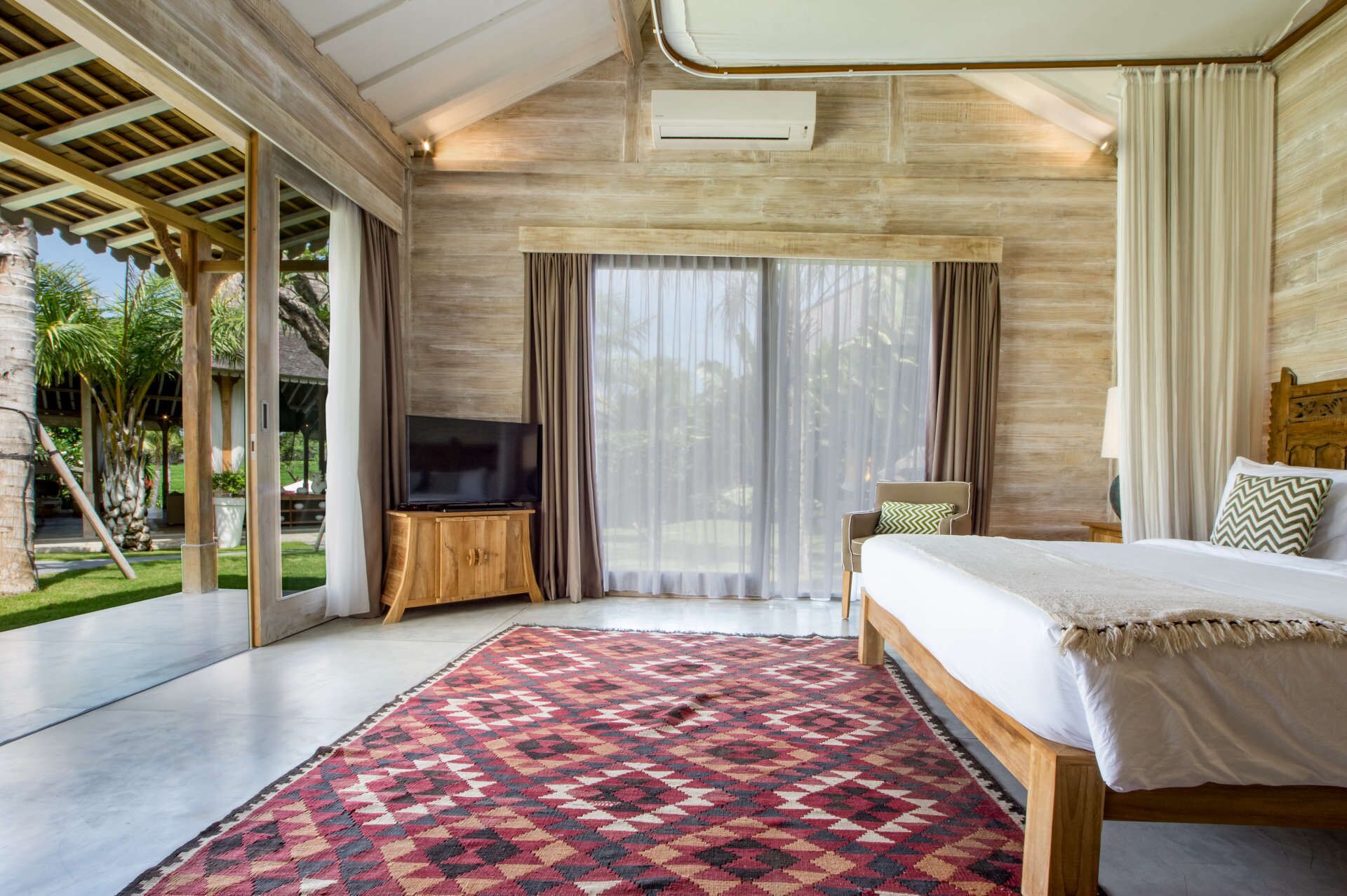 Un dormitorio amplio con una cama grande, un televisor sobre un soporte de madera, y vistas a un jardín exuberante a través de puertas correderas.