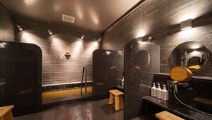 A modern bathroom with dark tiled walls, a wooden bench, a sink with a mirror, and a shower area. - Super Hotel Premier Saitama Omiyaeki Higashiguchi (Saitama)