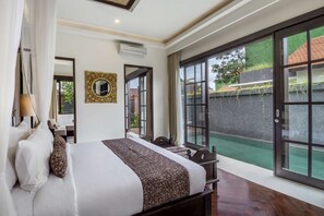 A bedroom with a large bed, a mirror, and a view of a green outdoor area through glass doors.